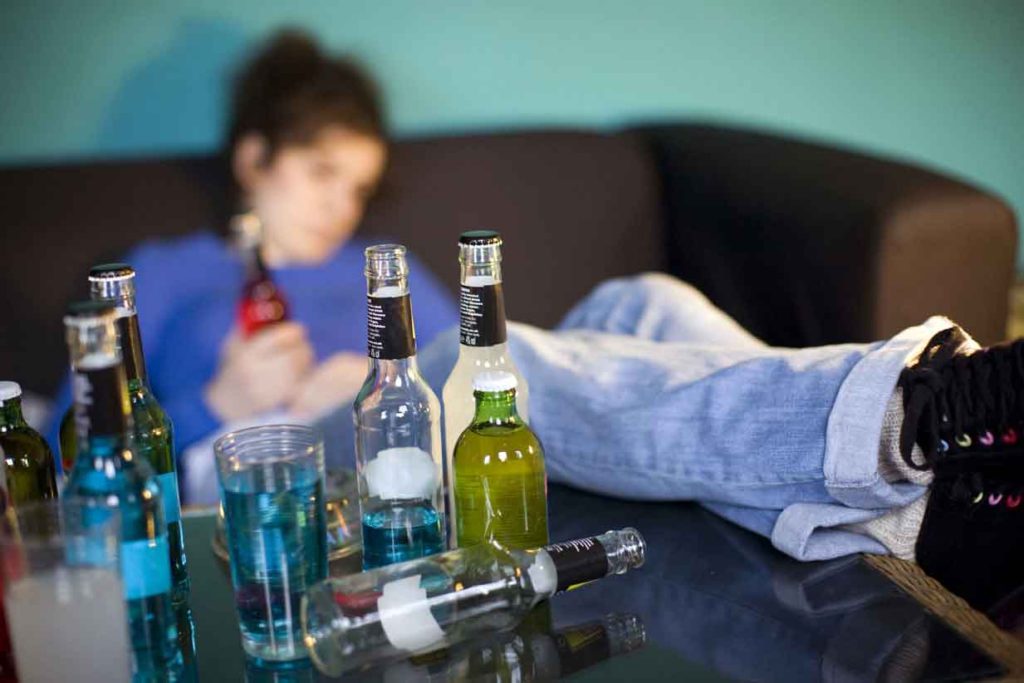 Consumo de alcohol en adolescentes. Joven sentado en un sofá rodeado de botellas de alcohol, representando el consumo problemático en adolescentes.