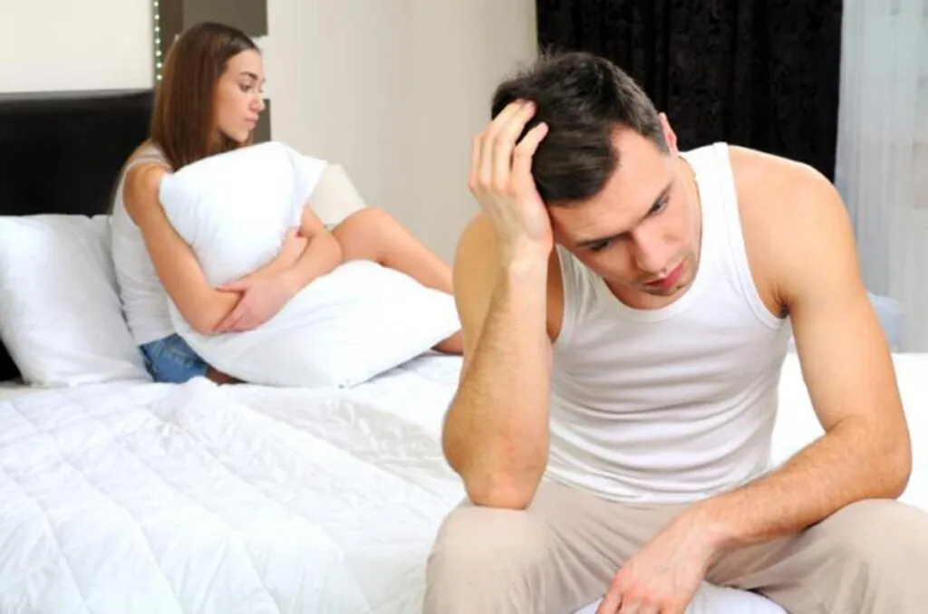 Hombre joven en la cama junto a una mujer, los dos con cara de preocupación