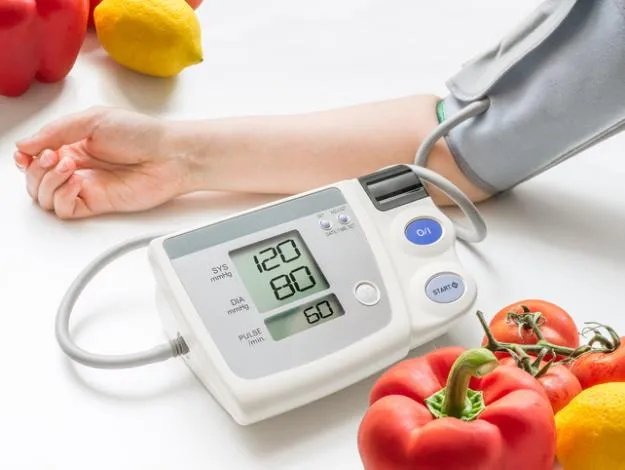 Brazo con baumanómetro digital marcando presión normal de 120/80 rodeado de vegetales frescos para controlar la hipertensión naturalmente