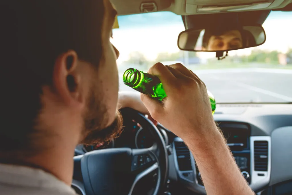 Conductor sosteniendo una botella de cerveza mientras maneja, simbolizando el riesgo de conducir bajo los efectos del alcohol.