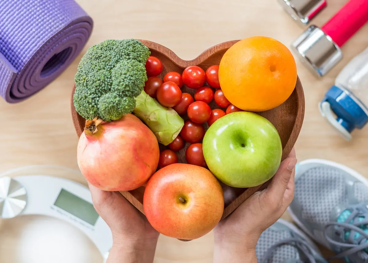 Manos sosteniendo un recipiente de frutas en forma de corazón rodeado de equipo de ejercicio, simbolizando la recuperación de la salud cardiovascular tras dejar el cigarrillo