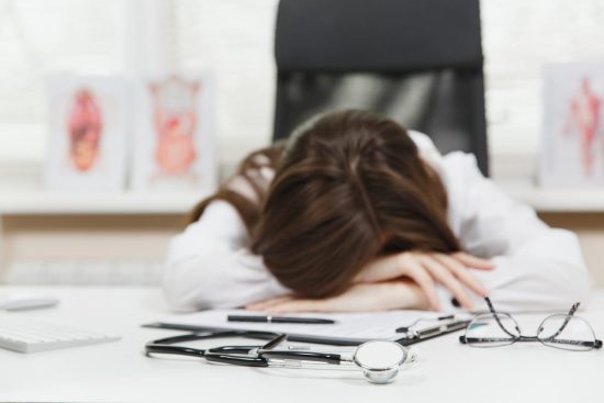 Estrés y agotamiento en los profesionales de la salud Médica agotada con la cabeza apoyada sobre el escritorio junto a su estetoscopio y documentos, reflejando el estrés y el cansancio en el ámbito sanitario.