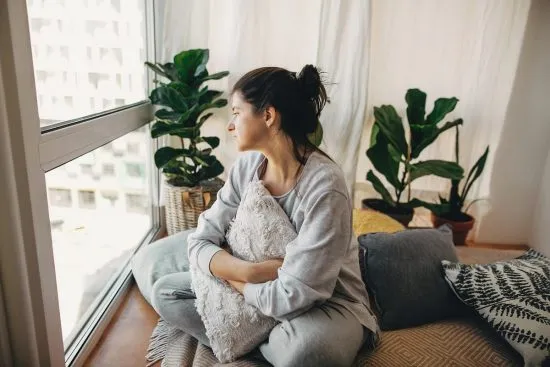 Mujer joven sentada en el suelo junto a la ventana abrazando un cojín con mirada melancólica, representando el tiempo de duelo necesario para sanar y procesar el dolor
