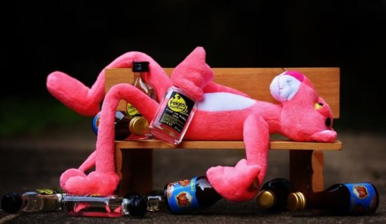 Peluche rosa recostado en un banco rodeado de botellas vacías, representando de forma humorística los efectos del exceso de alcohol.