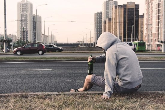 Joven con sudadera gris sentado al borde de una calle sosteniendo una botella de cerveza, representando consumo de alcohol en la vía pública.